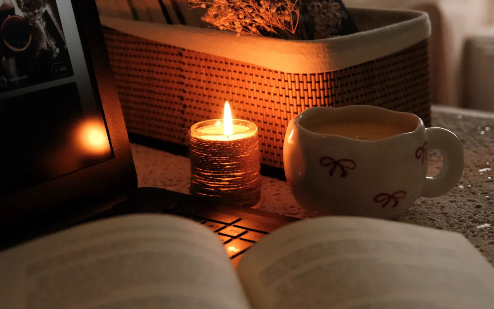A cup of coffee and a lit candle beside an open book and a glowing laptop keyboard.