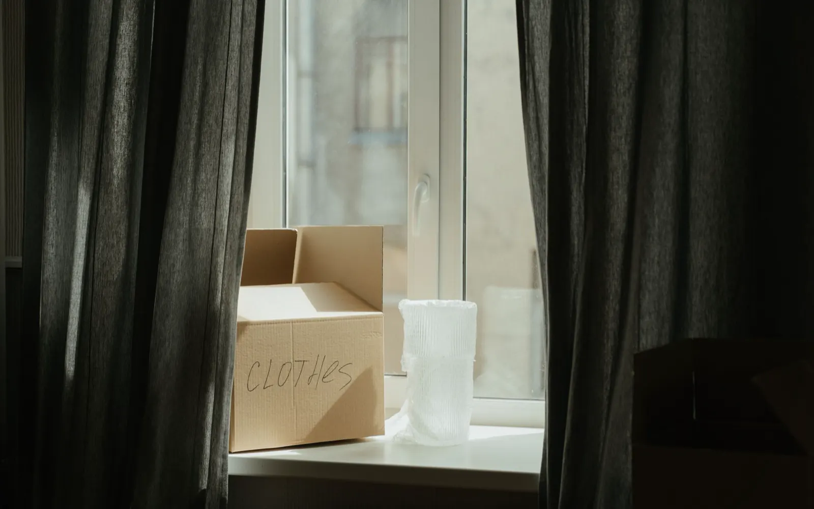 A packed moving box marked clothes on a windowsill between dark curtains.