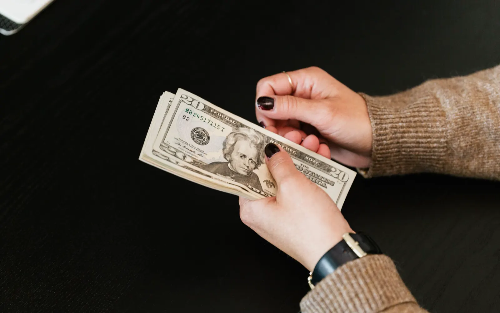 Two hands counting a stack of twenty-dollar bills over a dark table.
